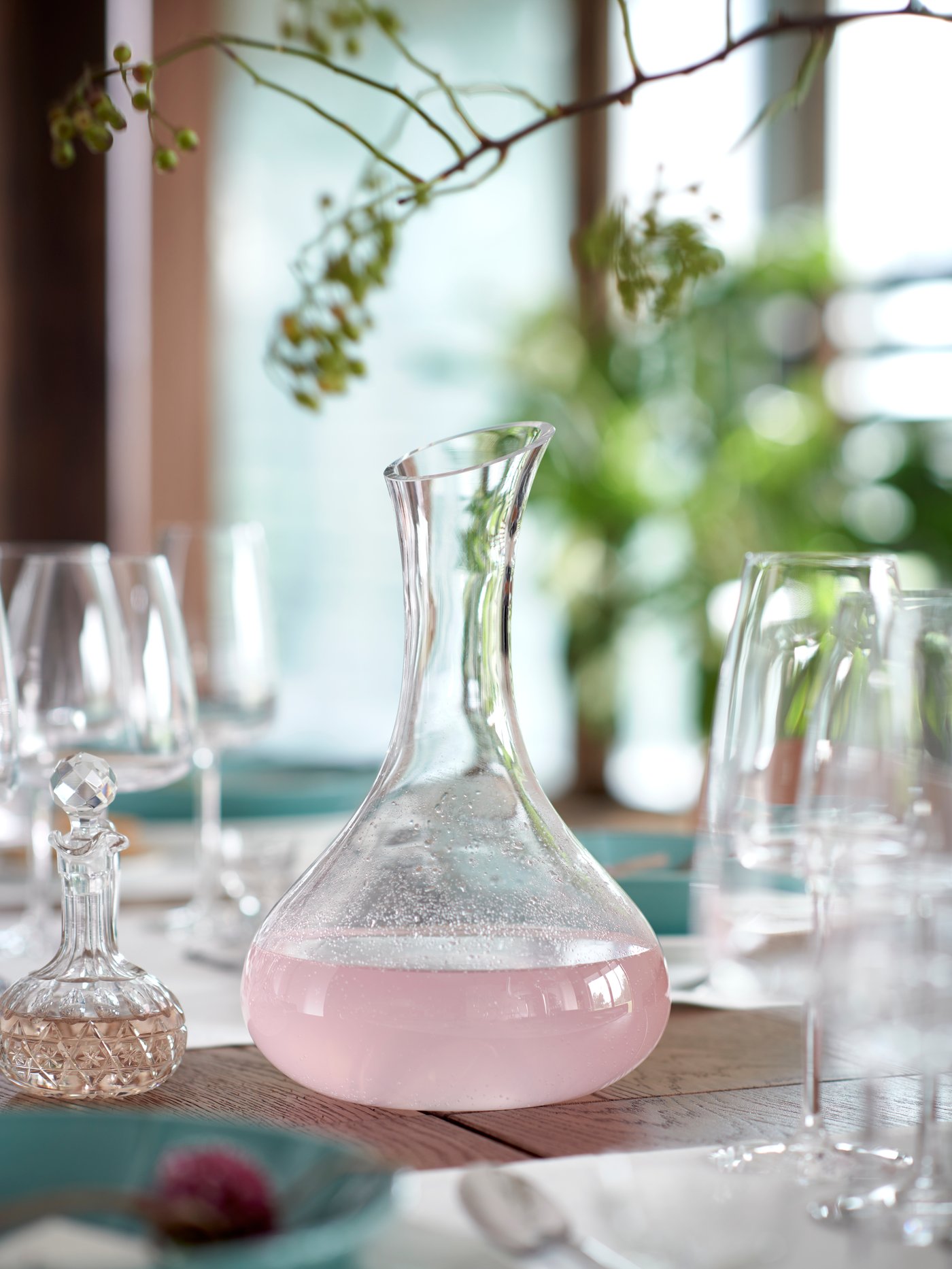 Sobre una mesa llena de vasos transparentes hay una jarra. Sobre ella cuelga una rama decorativa.
