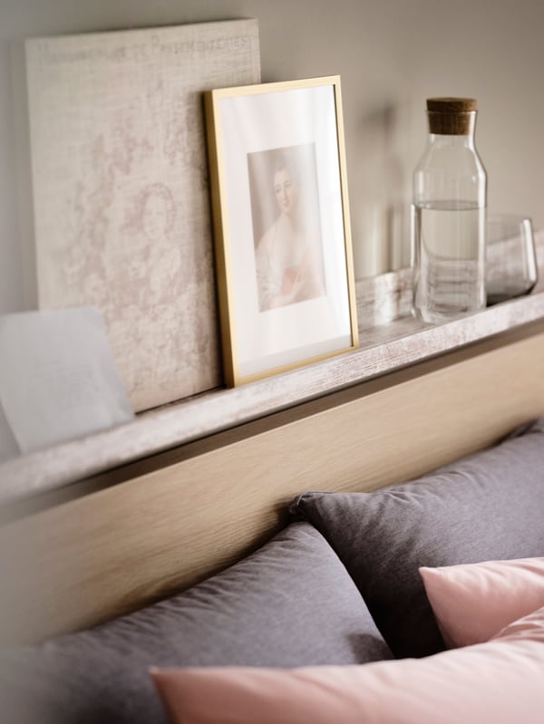 A MOSSLANDA picture ledge with framed pictures and a glass jar standing on it, above a bed with pillows.