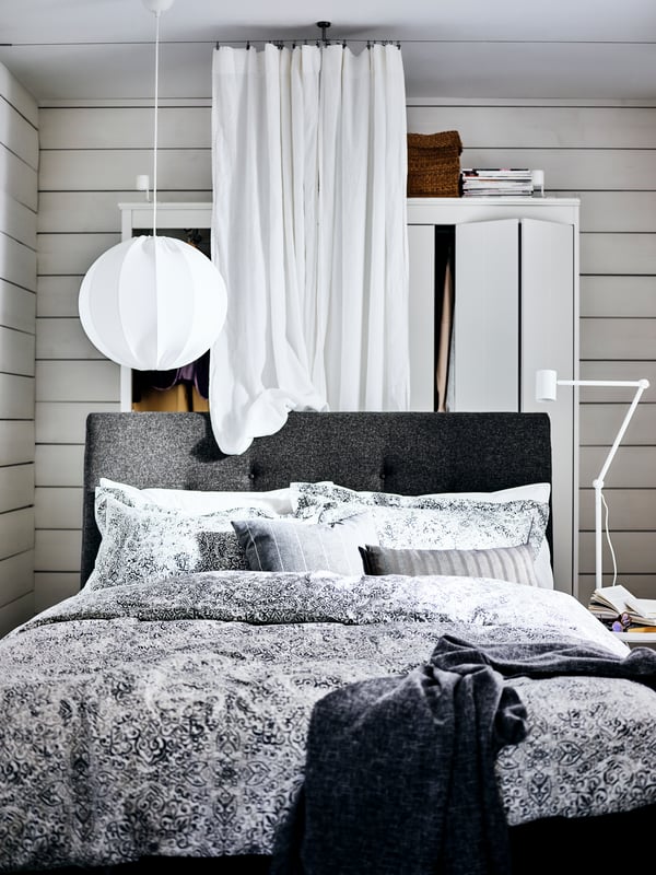 Un lit matelassé IDANÄS gris foncé avec une armoire et un rideau blanc derrière le lit, et une liseuse NYMÅNE à côté.
