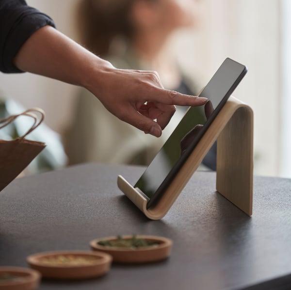 A person scrolls a tablet on a bamboo veneer tablet stand on a black worktop, plus another person sitting by a window.