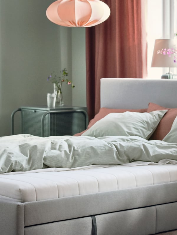 Katil upholsteri GLADSTAD dengan kotak storan dan tilam busa ÅSVANG diletakkan di bilik tidur berdekatan tingkap.