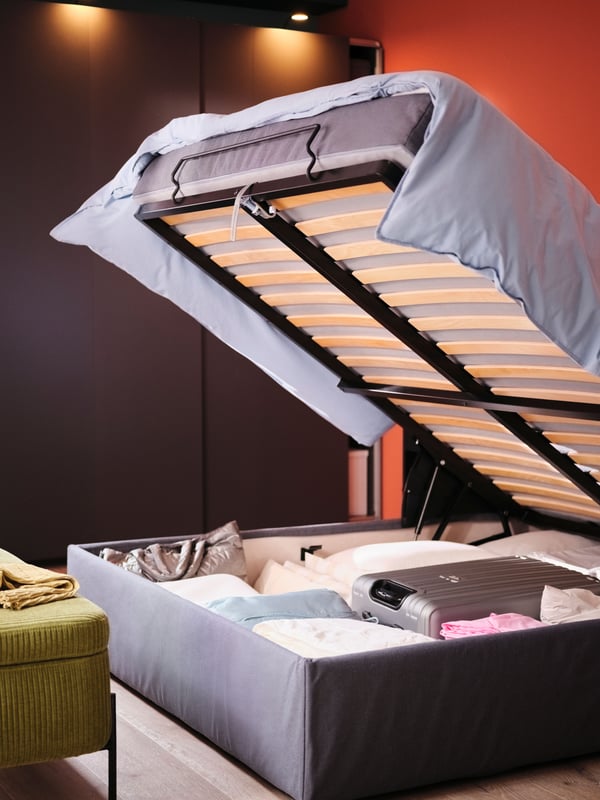 Lit coffre/sommier BLÅFJÄLLET dont la partie supérieure est relevée, révélant un espace de rangement accueillant une valise, du linge de lit et d’autres affaires.