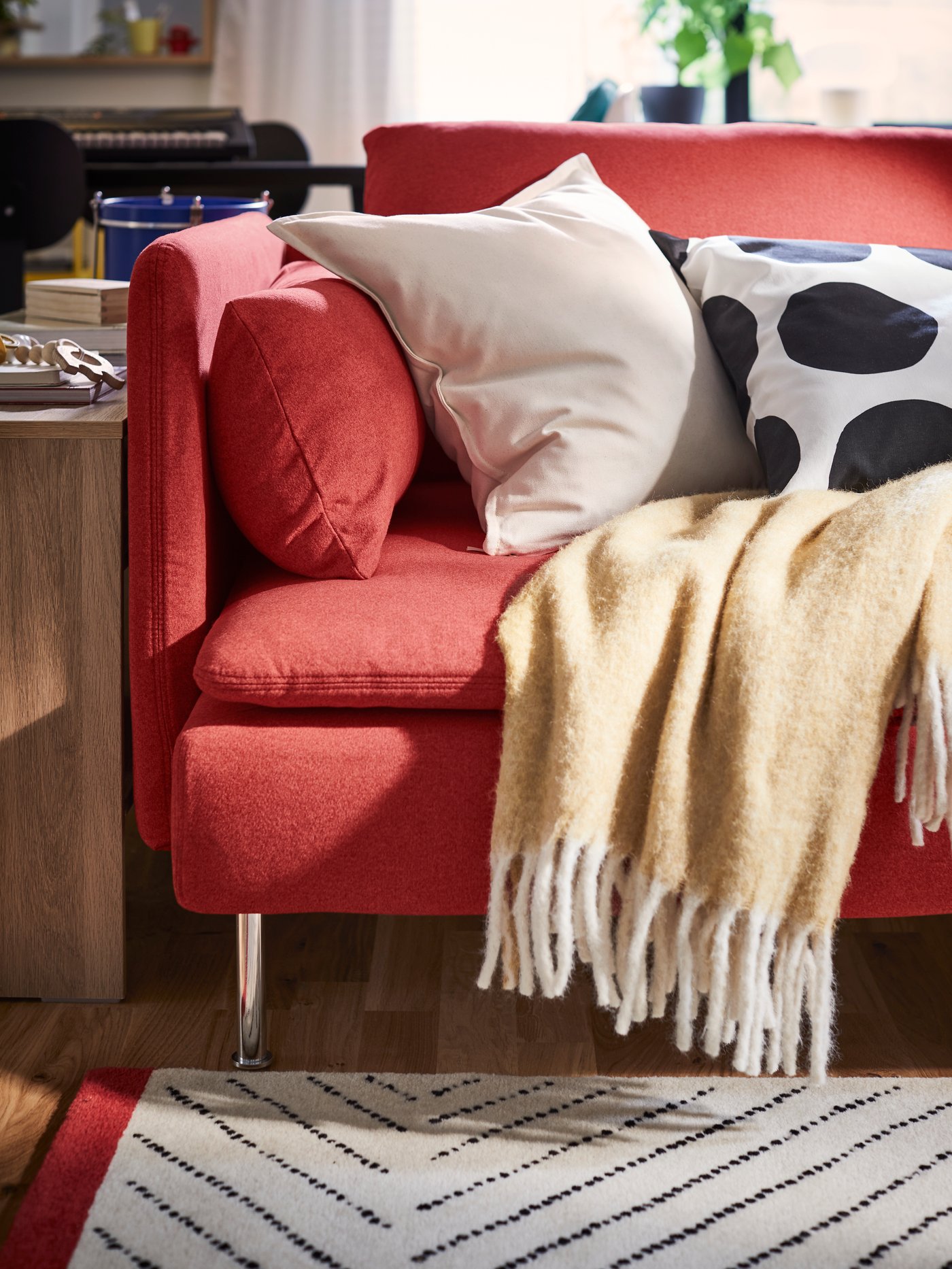 A beige-yellow KLIPPOXEL rug and two extra pillows lie on a red SÖDERHAMN sofa in a room with an UNDERVISNING rug.