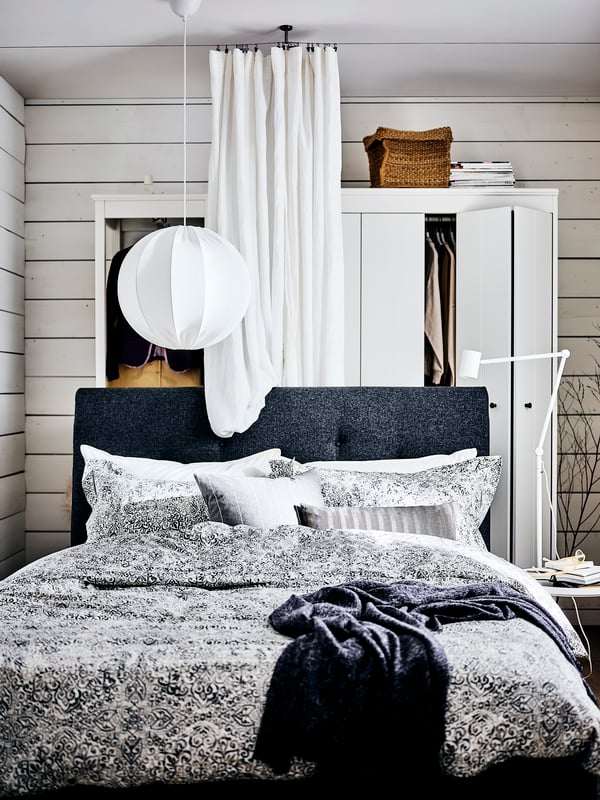 A dark gray IDANÄS upholstered bed covered in ÄNGSKLOCKA bed linen, in front of a white curtain and two wardrobes.