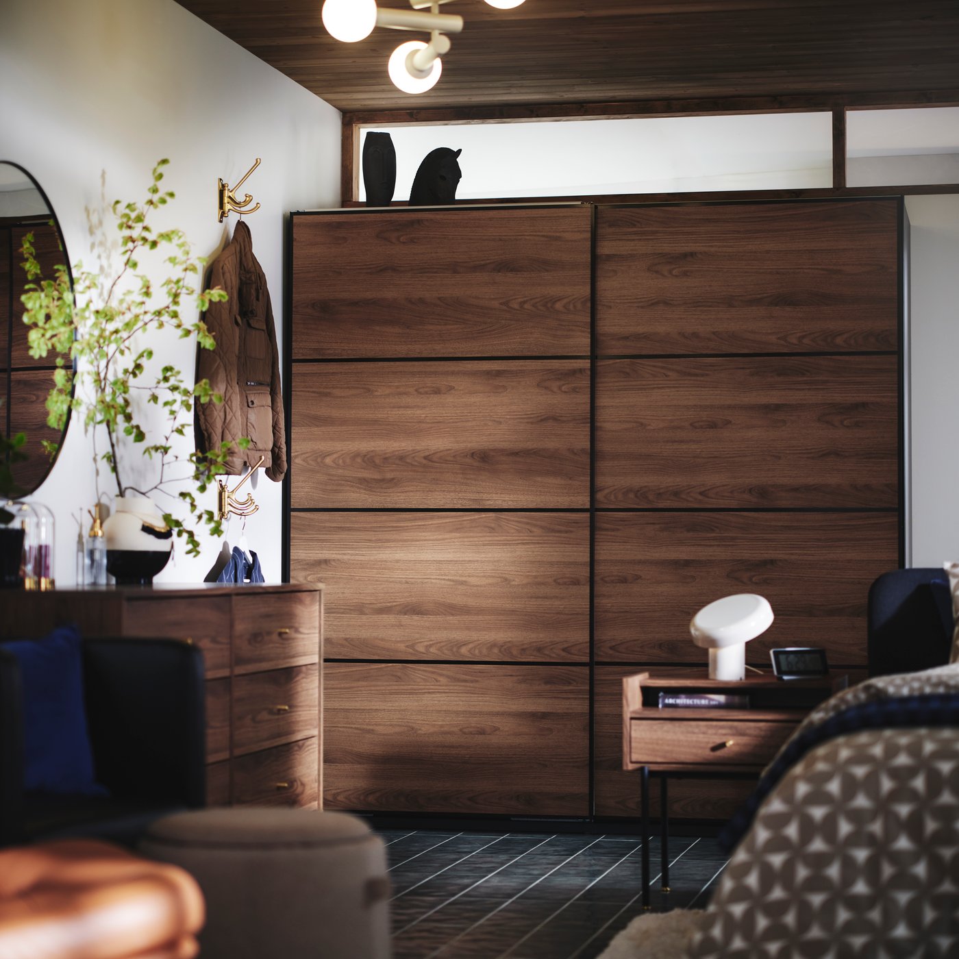 The side of a bed, RÅDMANSÖ bedside table and PAX MEHAMN wardrobe in dark wood and sliding doors in the background.