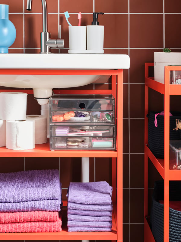 A wash-basin with a red-orange base frame, light red/purple towels, a transparent mini chest of drawers and toilet paper.