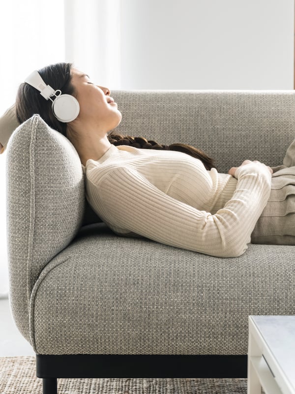 En kvinna med vita hörlurar som ligger och lyssnar på musik i en beige soffa med svarta ben