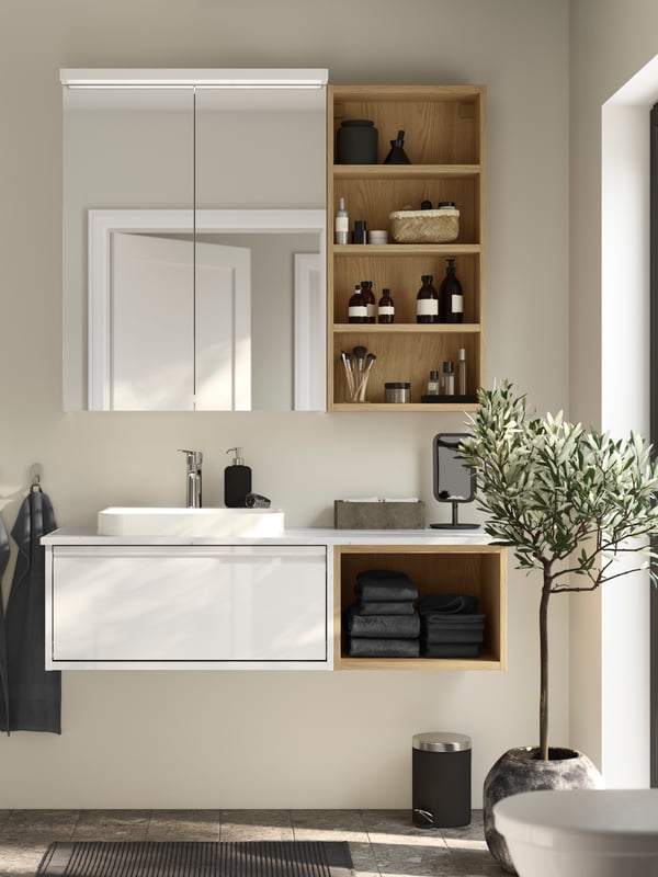Une armoire à pharmacie blanche au-dessus d’un meuble lavabo ÄNGSJÖN doté d’un tiroir blanc très brillant et d’une étagère ouverte effet chêne.