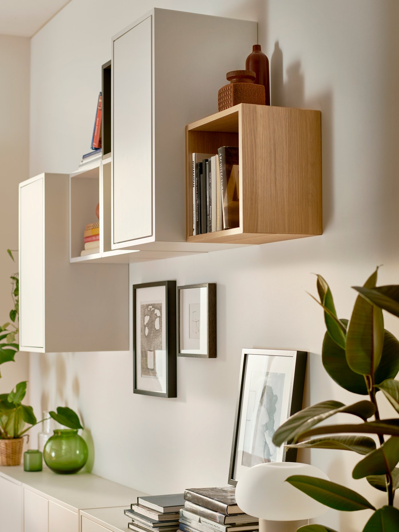 Una serie de repisas modulares EKET montadas en una pared, algunas blancas otras color madera. Debajo de ellas se aprecia un mueble con libros, cuadros y plantas.