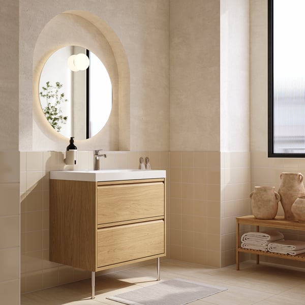 An oak effect, two-drawer ÄNGSJÖN/BACKSJÖN wash-stand with legs is below a round mirror in a modern beige and white bathroom.