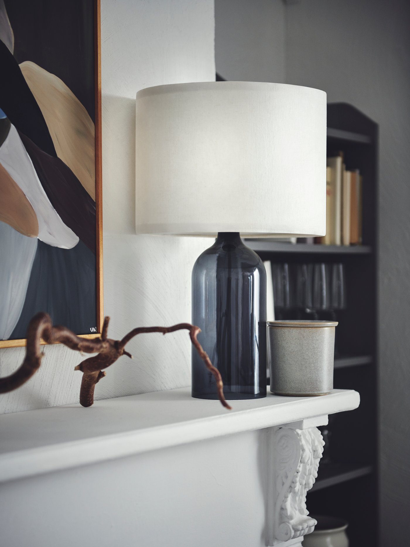 A smoked-glass-and-white TONVIS table lamp placed next to framed art on a mantlepiece in a light room.