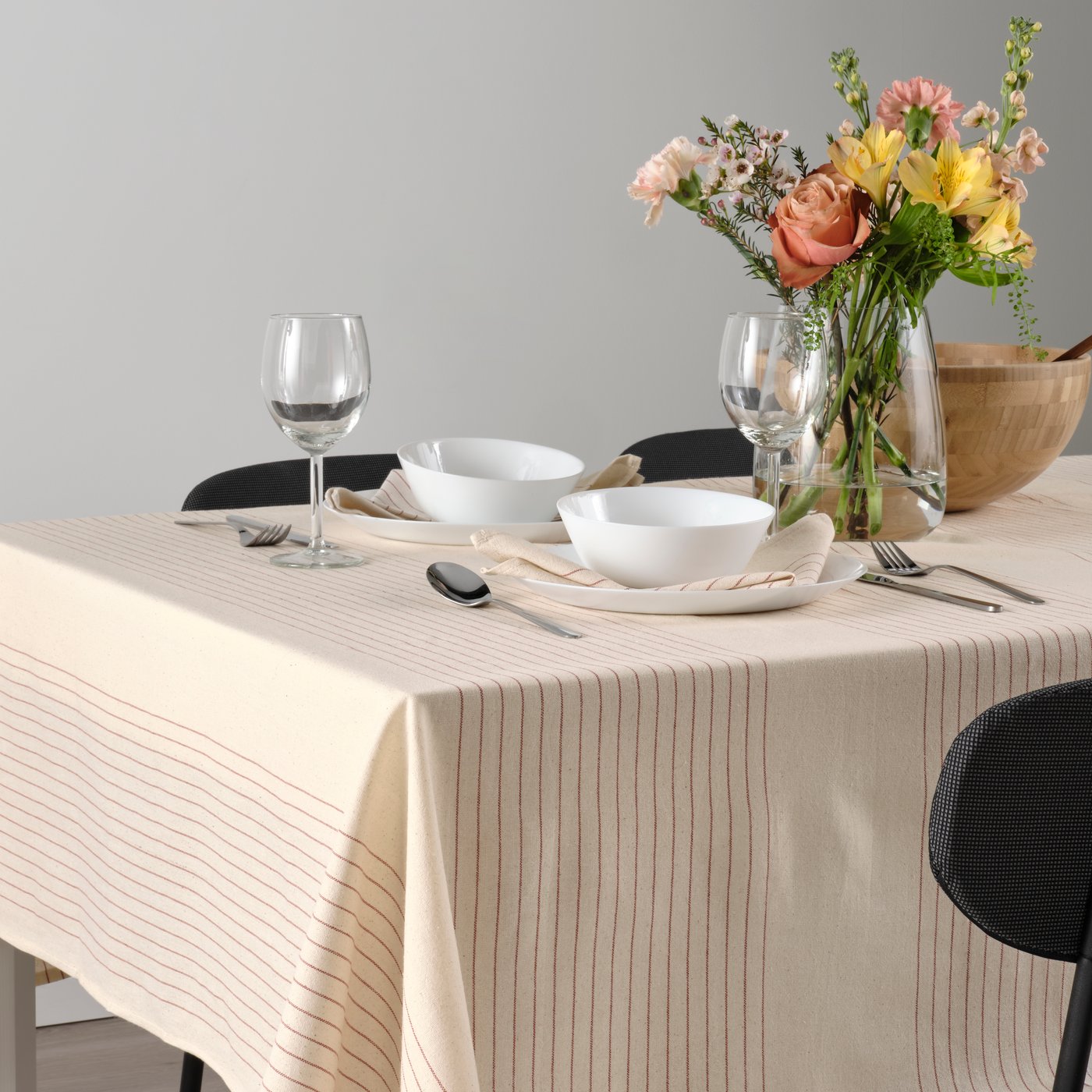 A dining table is dressed with a stripe pattern red/natural VIPPSTARR tablecloth, dinnerware and a vase with an artificial flower bouquet.