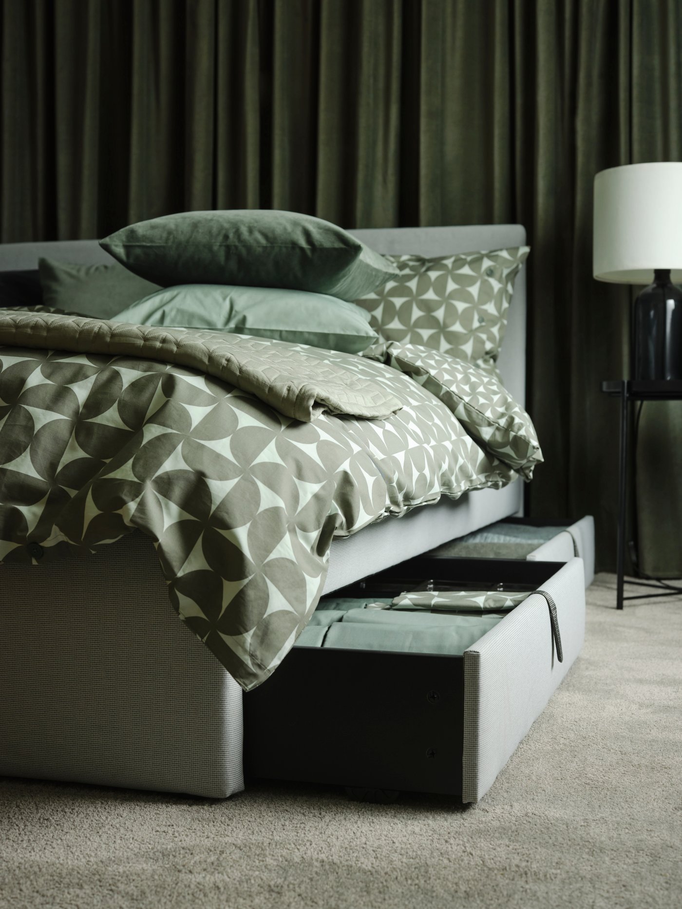 In a dark green room is a bed with green patterned bed linen. Drawers under the bed are pulled out to show folded linen.
