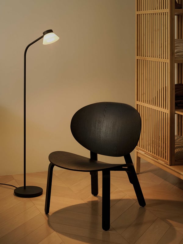 Svart FRÖSET träfåtölj med rund rygg och sits på trägolv, bredvid en svart golvlampa och en garderob i trä i bakgrunden