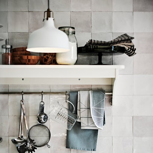A IDEALISK strainer and other baking tools hang on hooks from an off-white TORNVIKEN wall shelf, with a KORKEN jars and more kitchen utensils and textiles on top.