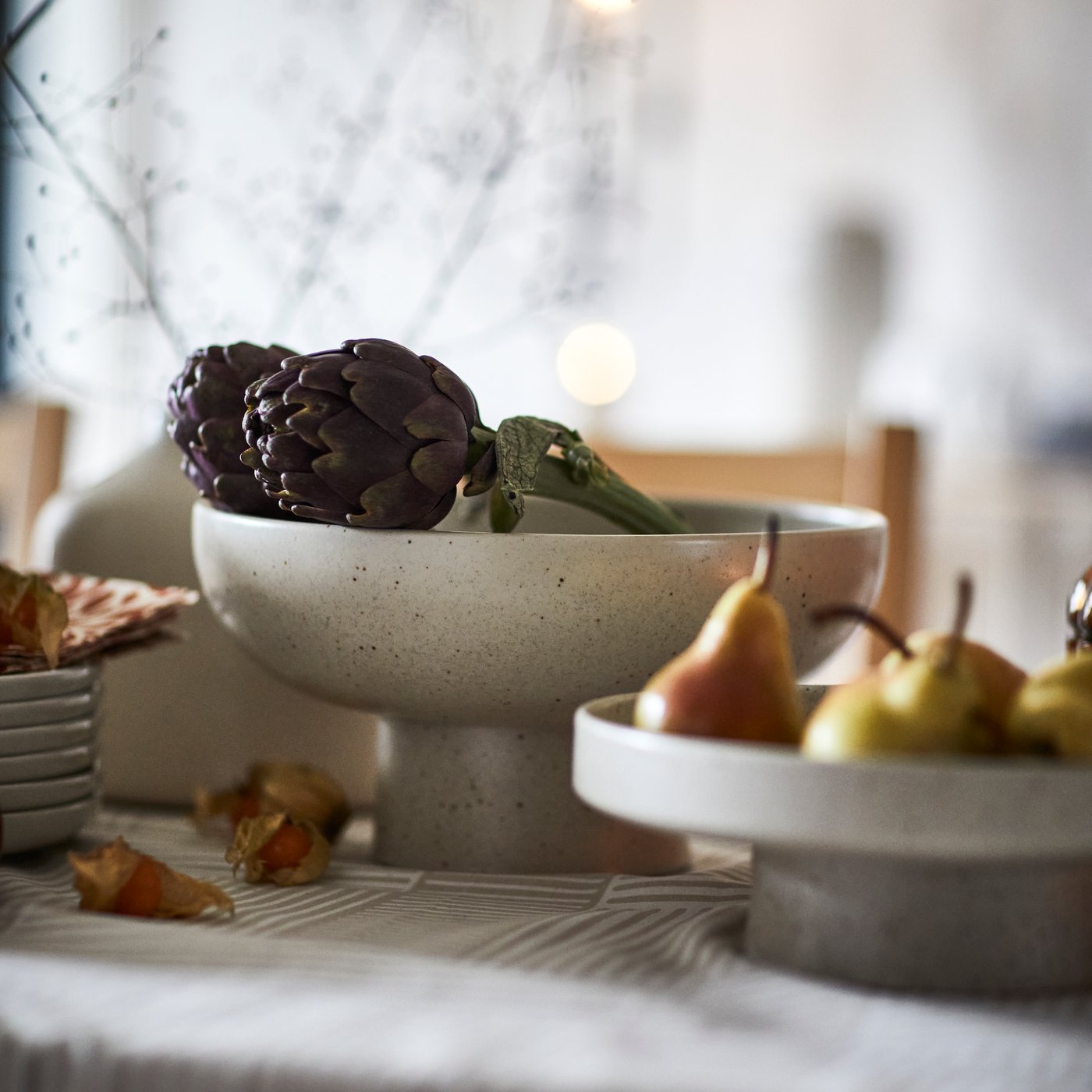 A light beige HÖSTAGILLE serving bowl holds artichokes on a table, next to a HÖSTAGILLE serving stand with pears in it.