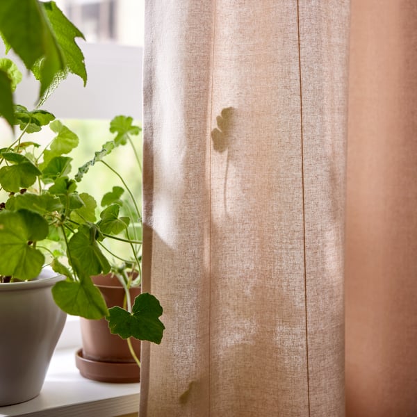 Two plant pots with plants stand on a windowsill with a light pink LENDA curtain hanging at one side of the window.