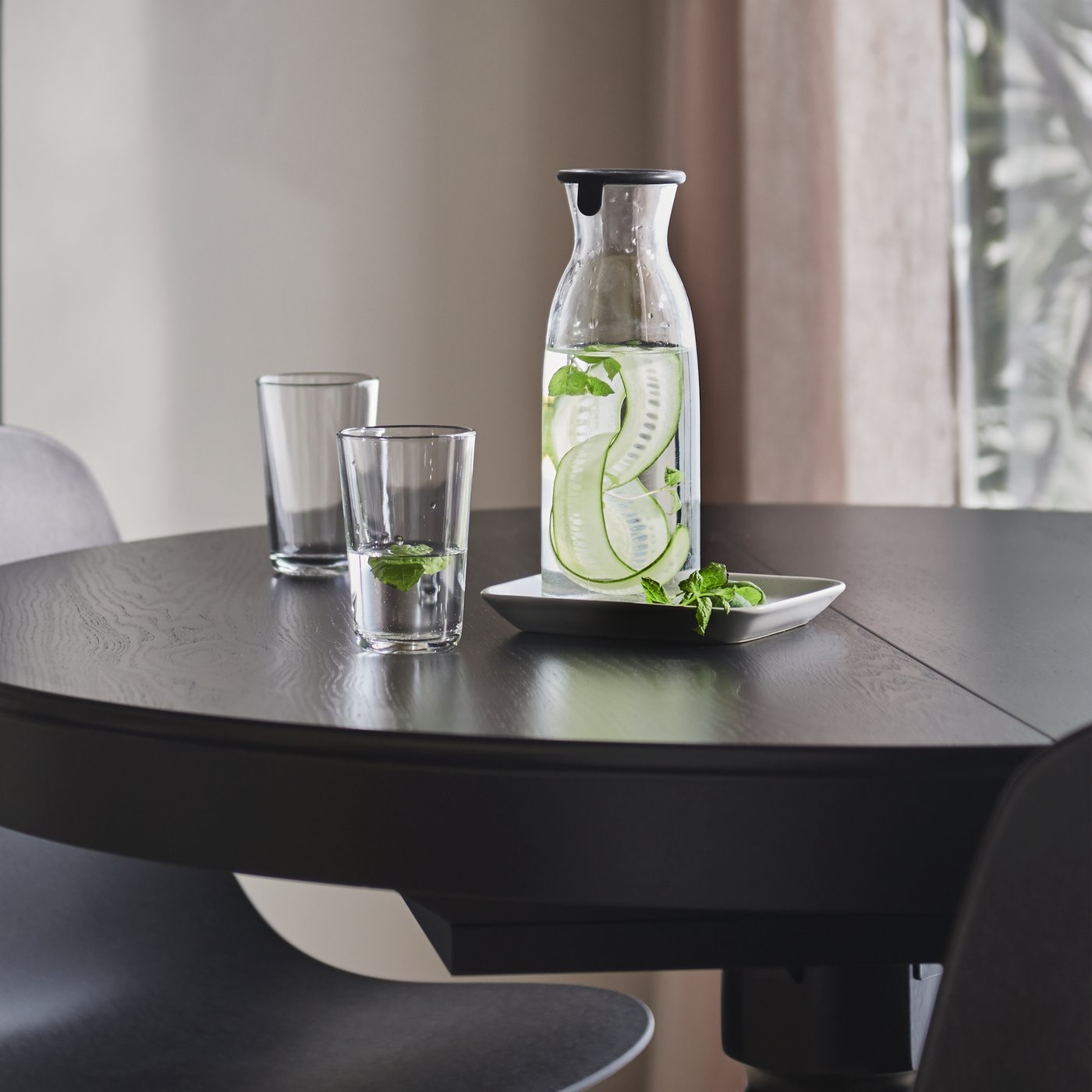 كأسان IKEA 365+ على طاولة مستديرة بلون أسود بجانب إبريق بغطاء به ماء وشرائح من الخيار.