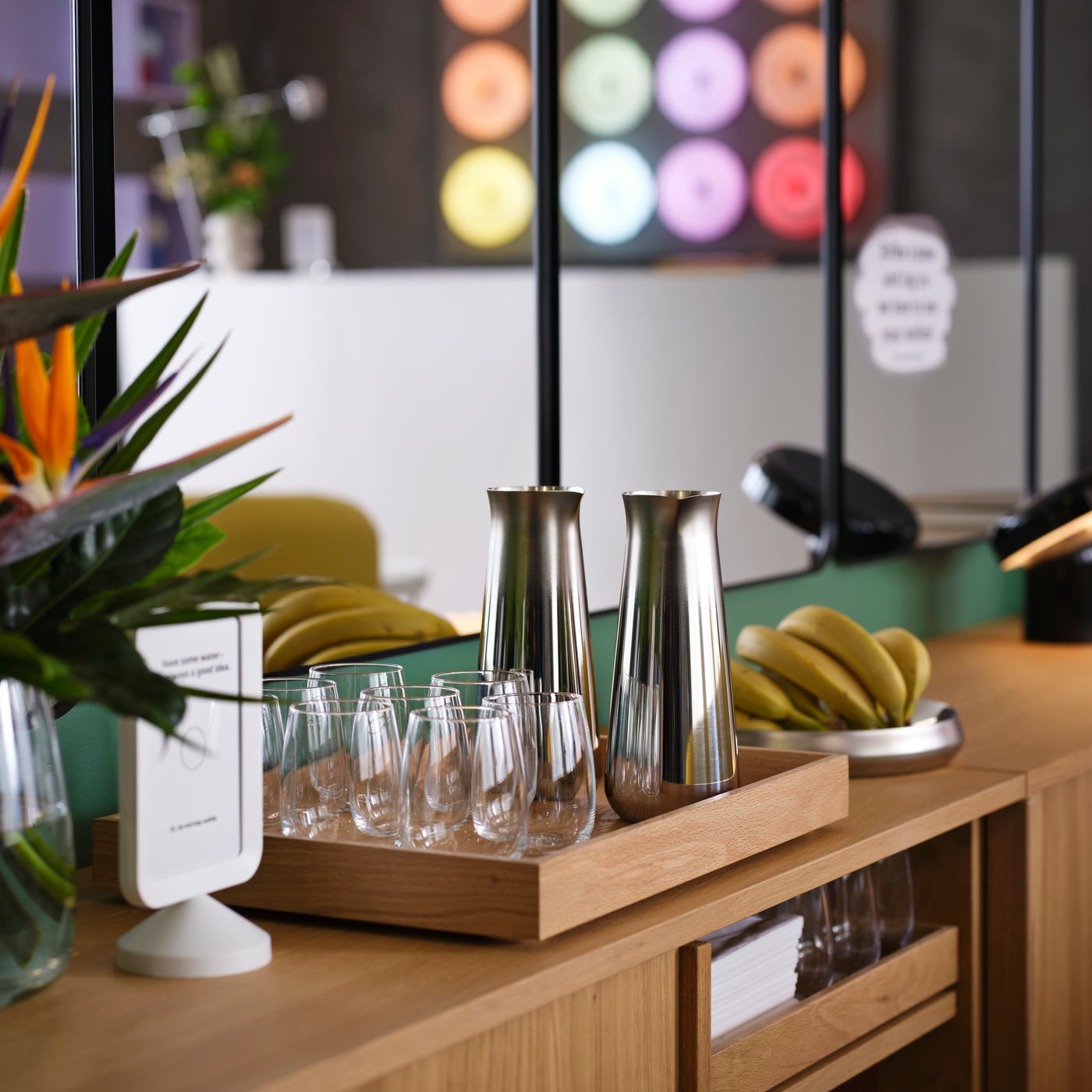 Two sleek stainless steel AFTONHAJ carafes are by drinking glasses and bananas on trays as a snack station in a hotel lounge.