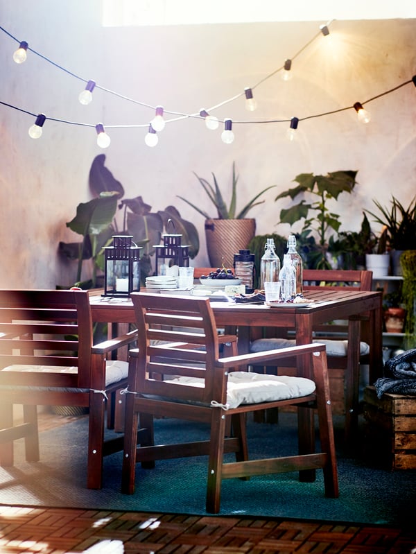 An outdoor table set with many chairs, lanterns, bottles and other items on the table and hanging lights above.