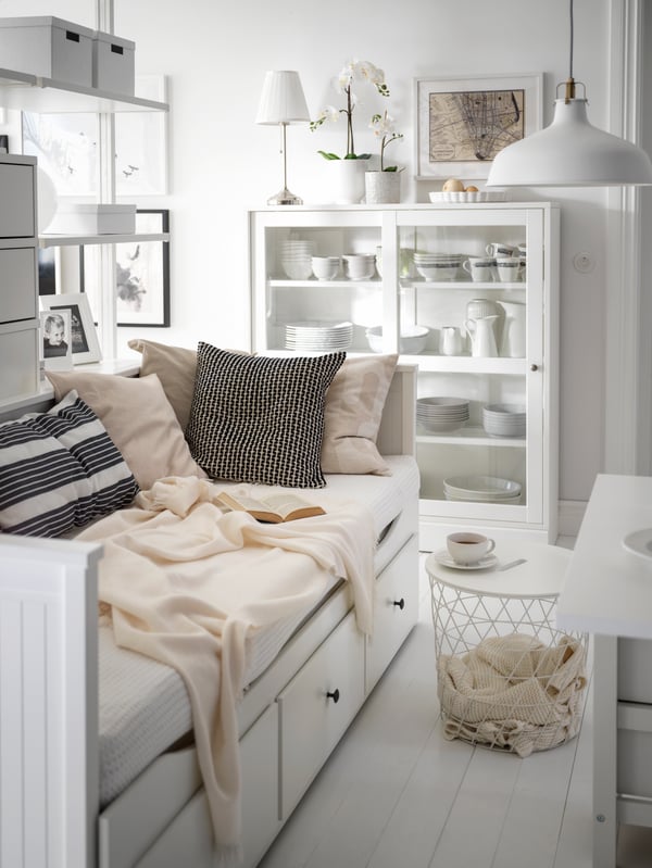 A white HEMNES day-bed laden with cushions and textiles, a white KVISTBRO storage table beside it, in a light living room.