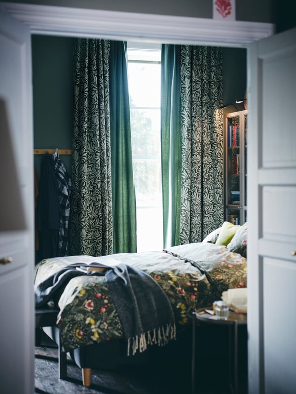 Une chambre avec des rideaux TRÅGSPINNARE vert foncé/motif feuilles et un lit avec une housse de couette et 2 taies KÄRRDUNÖRT.