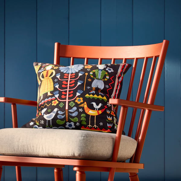 A multicolour TRATTKAKTUS cushion cover featuring vibrant floral and bird designs, displayed on a brown-red PERSBOL armchair.