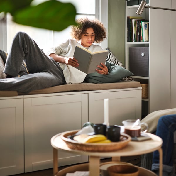 A person lies reading a book on a PLATSA storage bench under a window near a birch veneer BORGEBY coffee table.