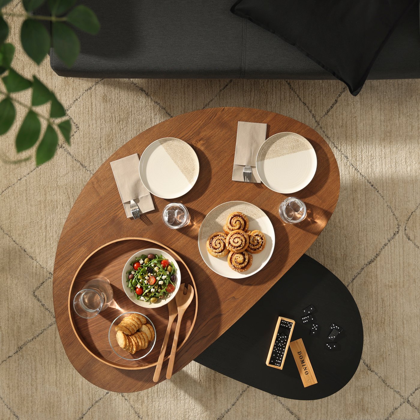 A top view shot of a ÖRTRÄSK nest tables in brown and black ash veneer placed on a rug next to a sofa in a living room setting.