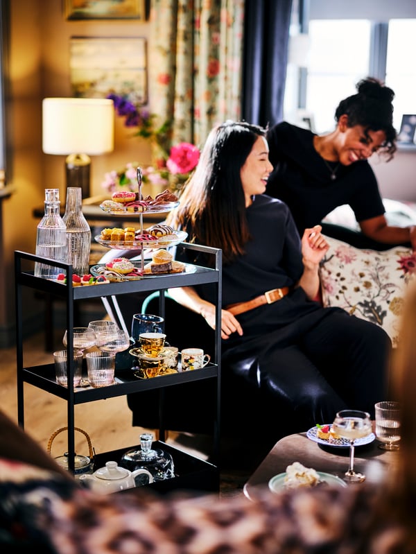 Two laughing women sitting next to a black NISSAFORS trolley filled with confections, glasses, teapots and teacups.