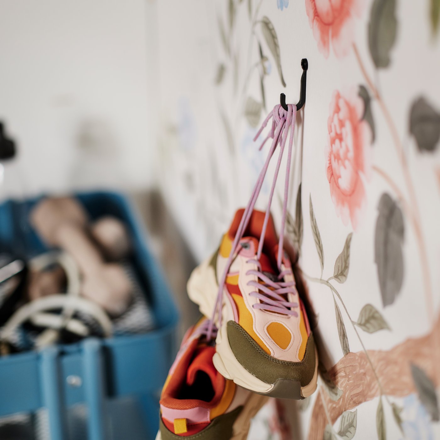 Crochet mural HÖGAFFEL fixé sur un mur couvert d’un papier peint fleuri ; des chaussures de sport colorées sont suspendues au crochet par leurs lacets.