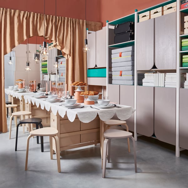 A set up dining table with a white table cloth, plates, cutlery and glasses is standing next to a storage unit.