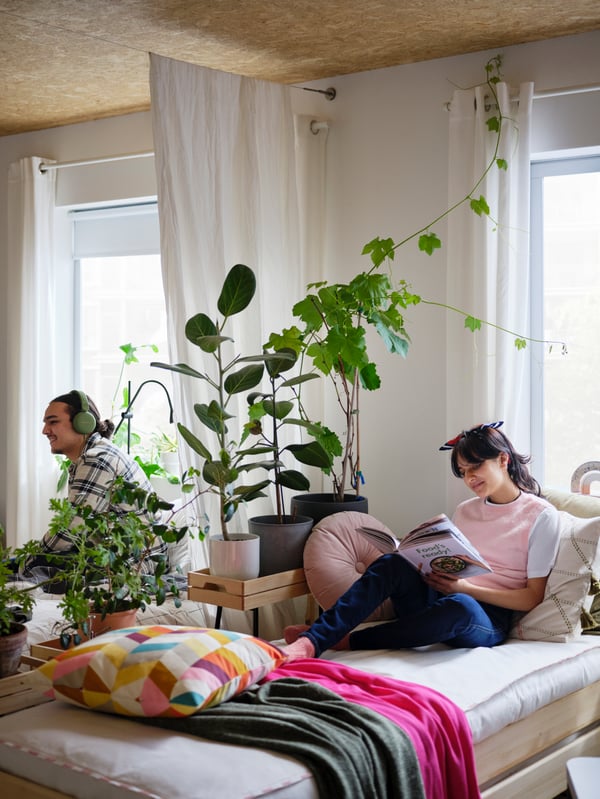 A woman on an UTÅKER stackable bed reads a book. Behind a room-dividing combo of a plant swath and a curtain sits a man.