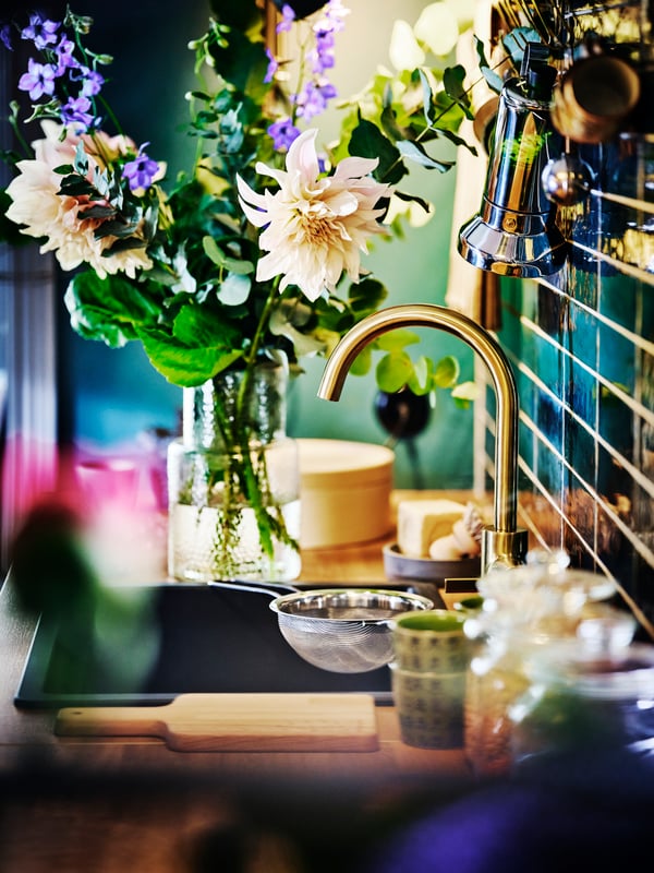 A kitchen sink with a brass DELSJÖN kitchen mixer tap against a blue-green tiled wall with a vase of flowers on the counter.
