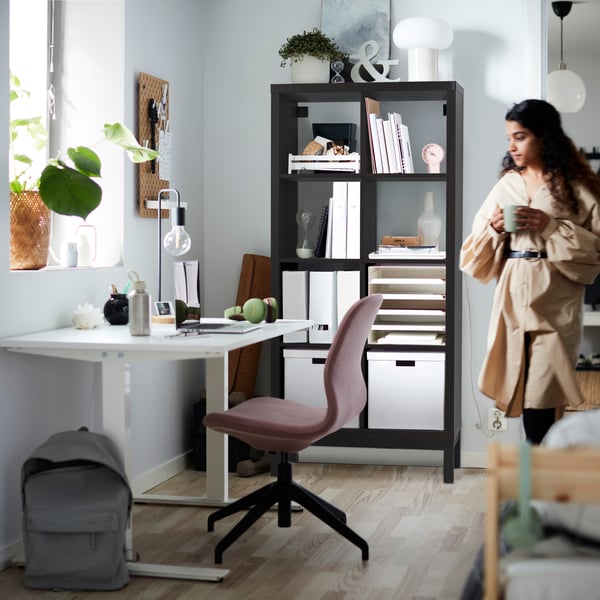 A teenage girl holding a mug stands in a room with a desk-and-chair combo and a black-brown KALLAX shelving unit.