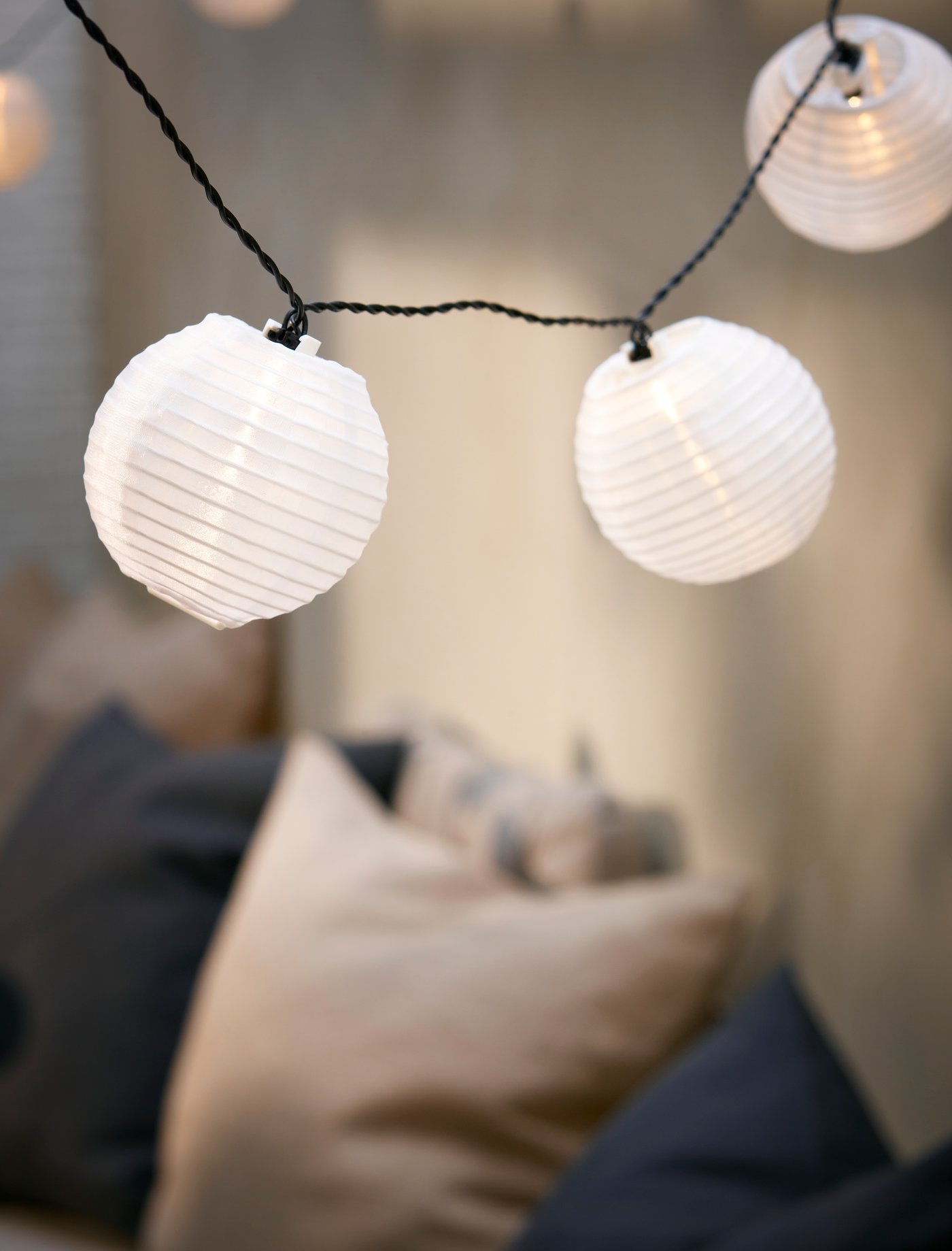 The white paper globes of a SOLARVET solar-powered LED lighting chain hang over a cluster of cushions in a dimly lit space.