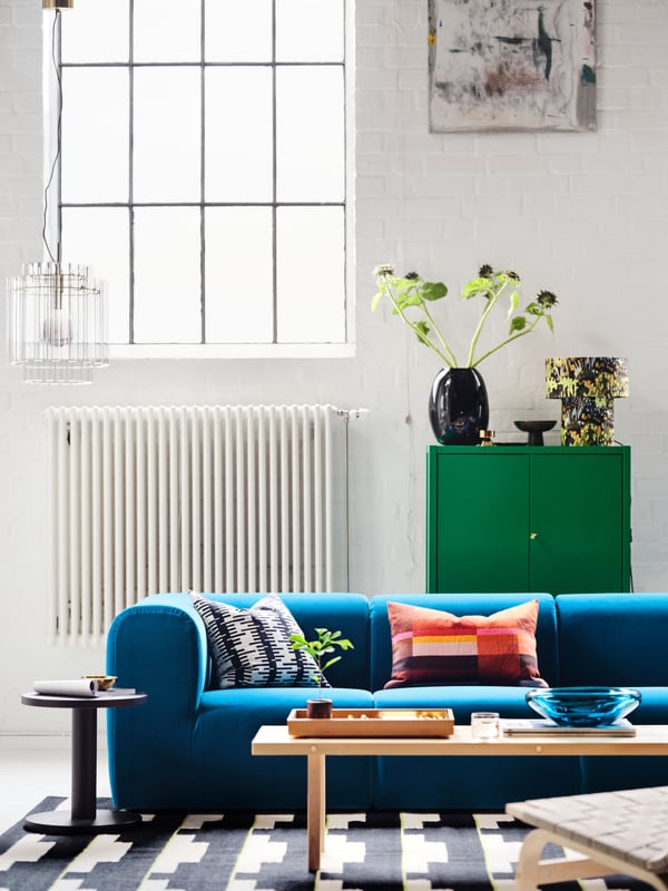 A room with a dark turquoise STOCKHOLM 2025 3-seat sofa, with two tables on a rug in front, and a green cabinet behind.