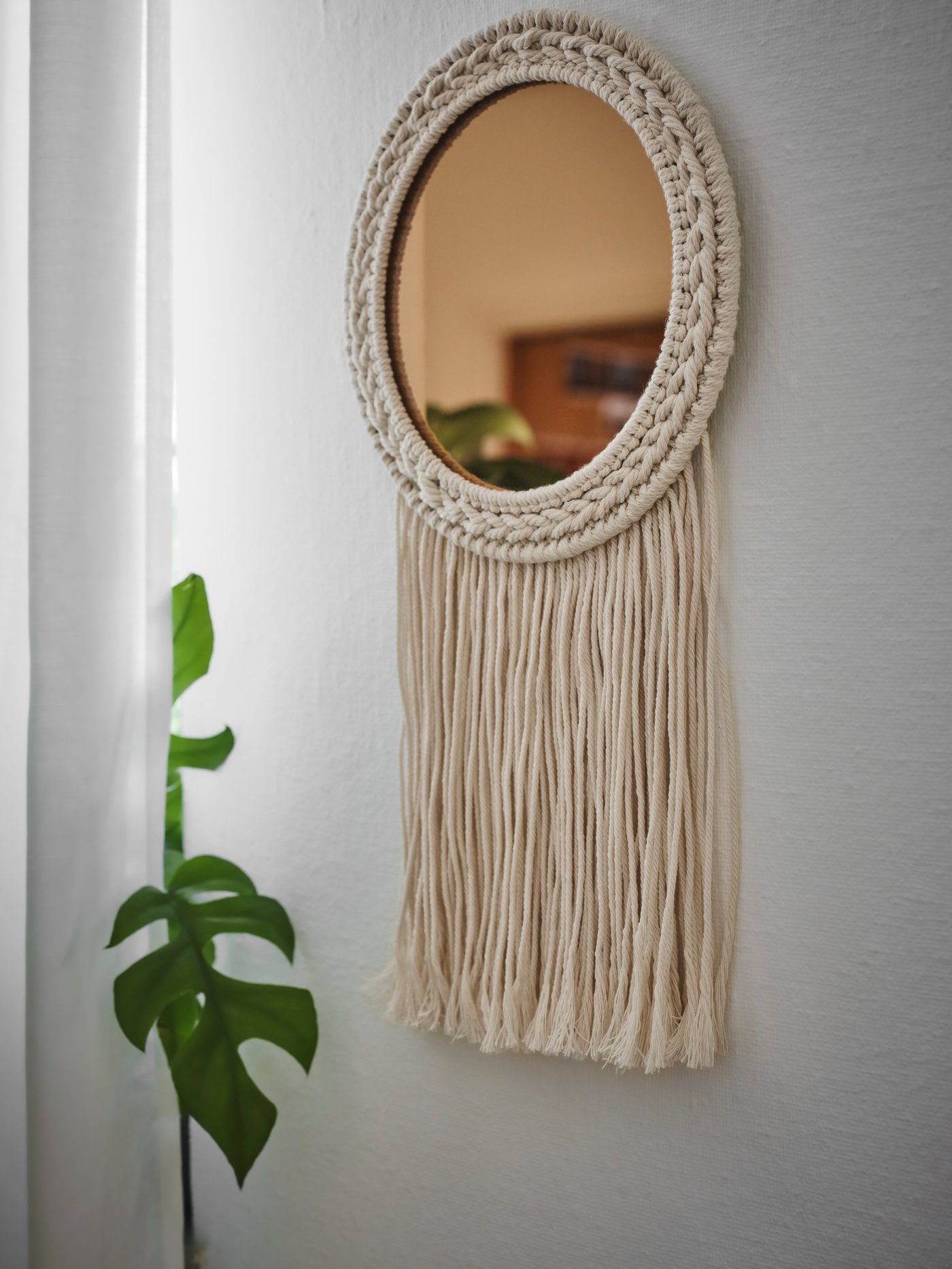 A beige copper-colour ENERGISKOG decorative mirror with fringes hangs on a wall near a large green plant and a curtain.