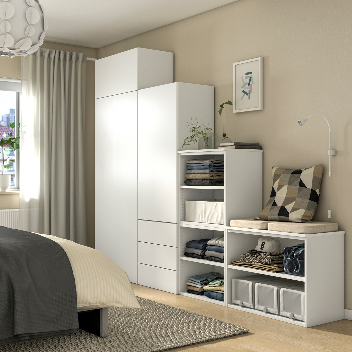 Modernes Schlafzimmer mit einem modularen, weißen Schranksystem aus verschiedenen Höhen und offenen sowie geschlossenen Fächern; daneben ein Bett, eine Nachttischlampe und dezente Wohnaccessoires.