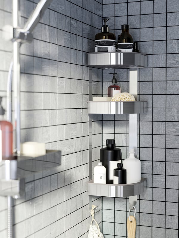 En la esquina de un espacio de ducha con los muros revestidos de mosaicos grises, se encuentra instalado a muro un estante esquinero BROGRUND de acero inoxidable, con botellas de shampoo, esponjas y jabones.
