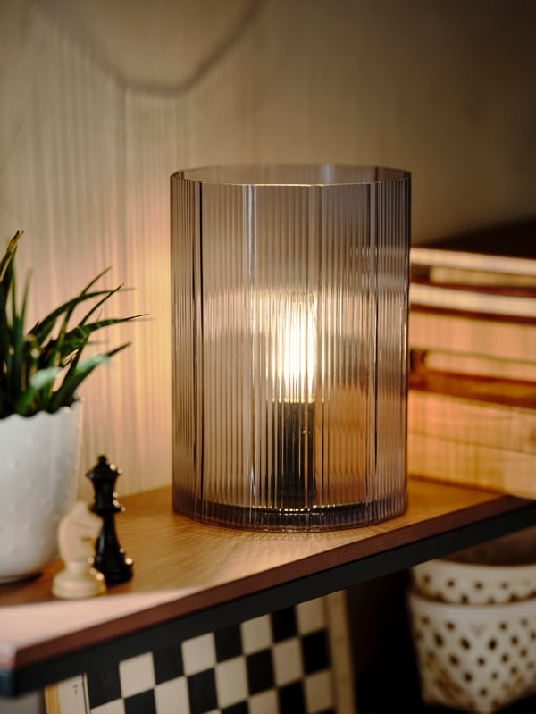 Sur une étagère murale, une lampe de table MIKROKLIN en verre bleu allumée installée parmi des livres, une plante en pot et des pièces d’échec.