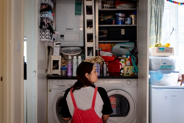 A lady crouches in front of her washing machine. Above there is a boiler and shelving stretching up to the ceiling, filled with lots of cleaning products and other items.