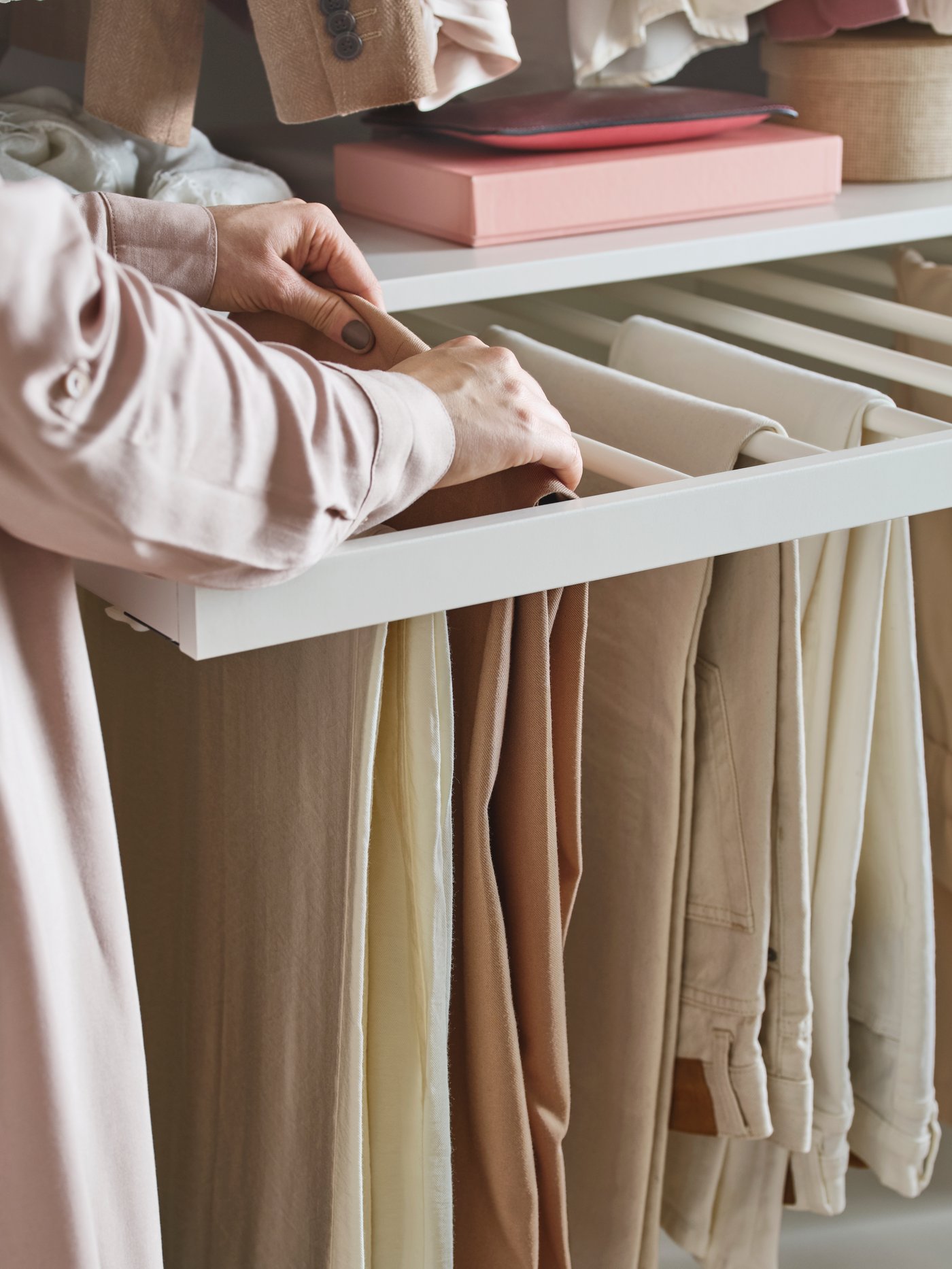 A women’s hands lift a pair of trousers out of a white KOMPLEMENT pull-out trouser hanger inside a PAX wardrobe.