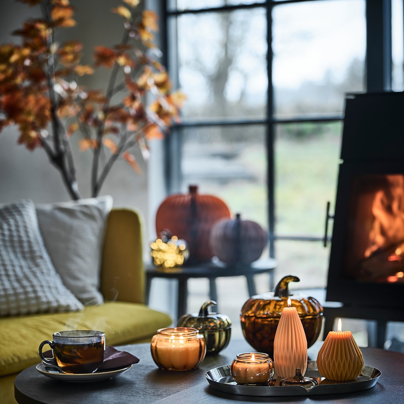 A cosy corner next to a window has a coffee table with lit HÖSTAGILLE scented candles, pumpkin-shaped pillar candles and mug.