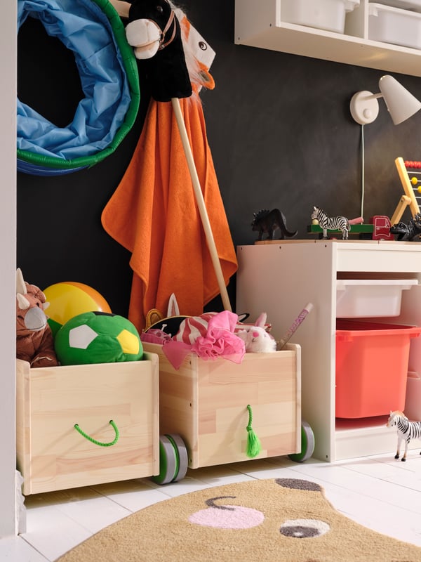 Zwei FLISAT Spielzeugwagen mit Spielzeug stehen vor einer Wand mit Tafelfarbe und neben einer TROFAST Kombination mit Boxen.