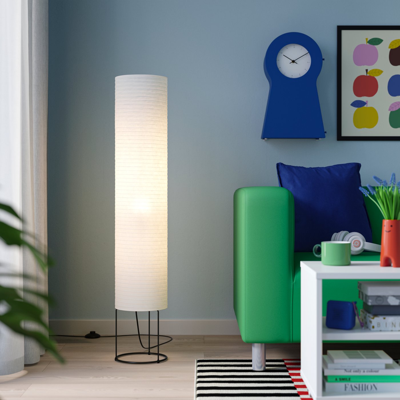 En lys stue med blå væg, grøn sofa, gulvlampe i rispapir, vægur, farverig vægkunst og et lille sofabord med pynt.