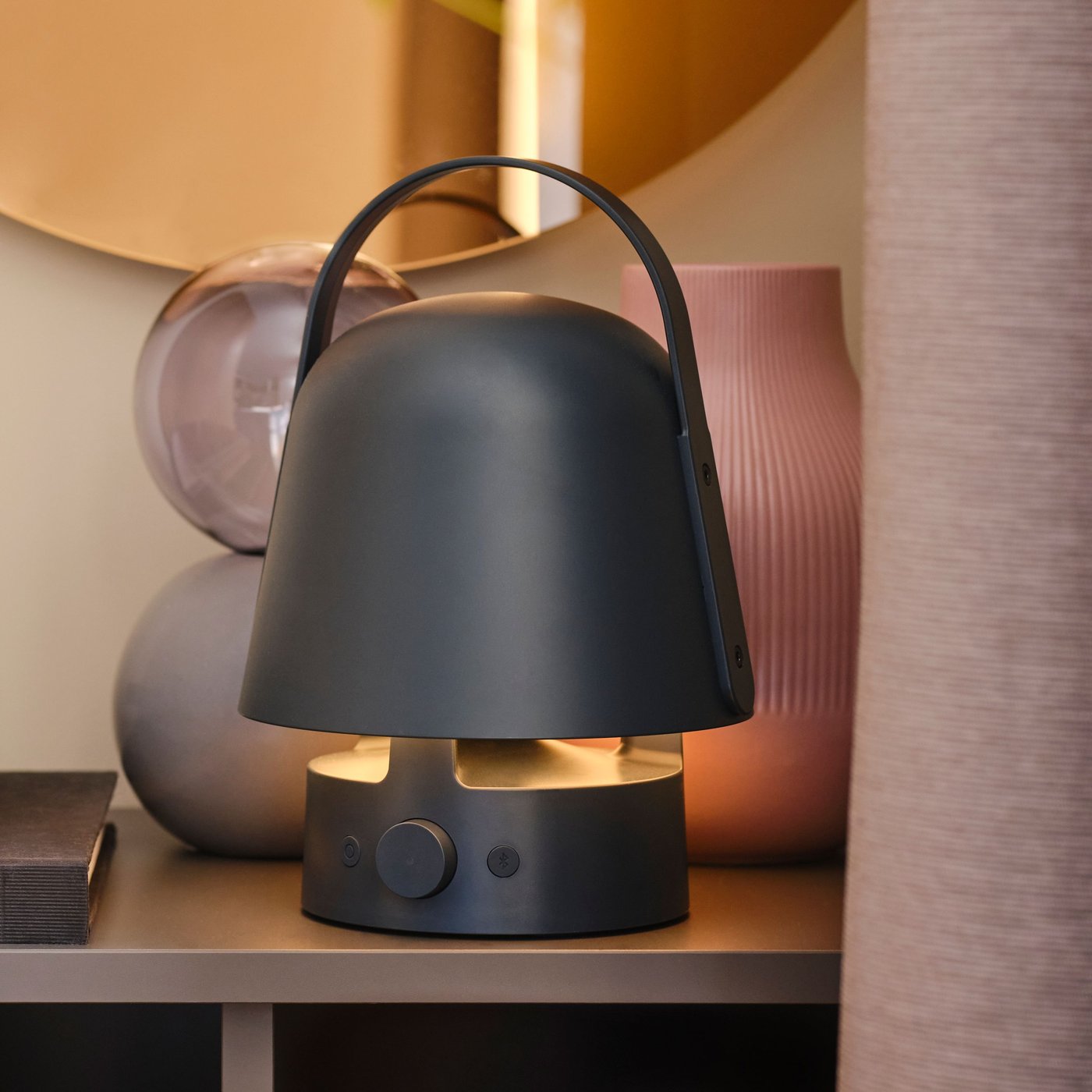 Applique/lampe LED VÄRMBLIXT ronde couleur bronze au-dessus d’un rangement où se trouvent des objets décoratifs et une lampe enceinte.