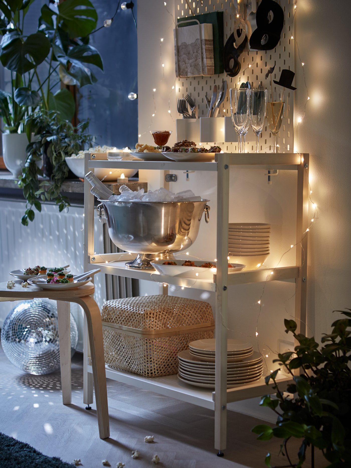 A white EKENABBEN open shelving unit holds plates, glasses, and a champagne bowl; fairy lights are strung around the unit.