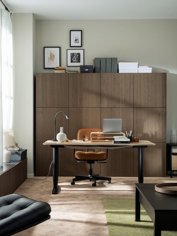 A softly sunlit, carpeted room with a tidy workspace, a wall of wood-front cabinets, a TRULSTORP coffee table and wall art.