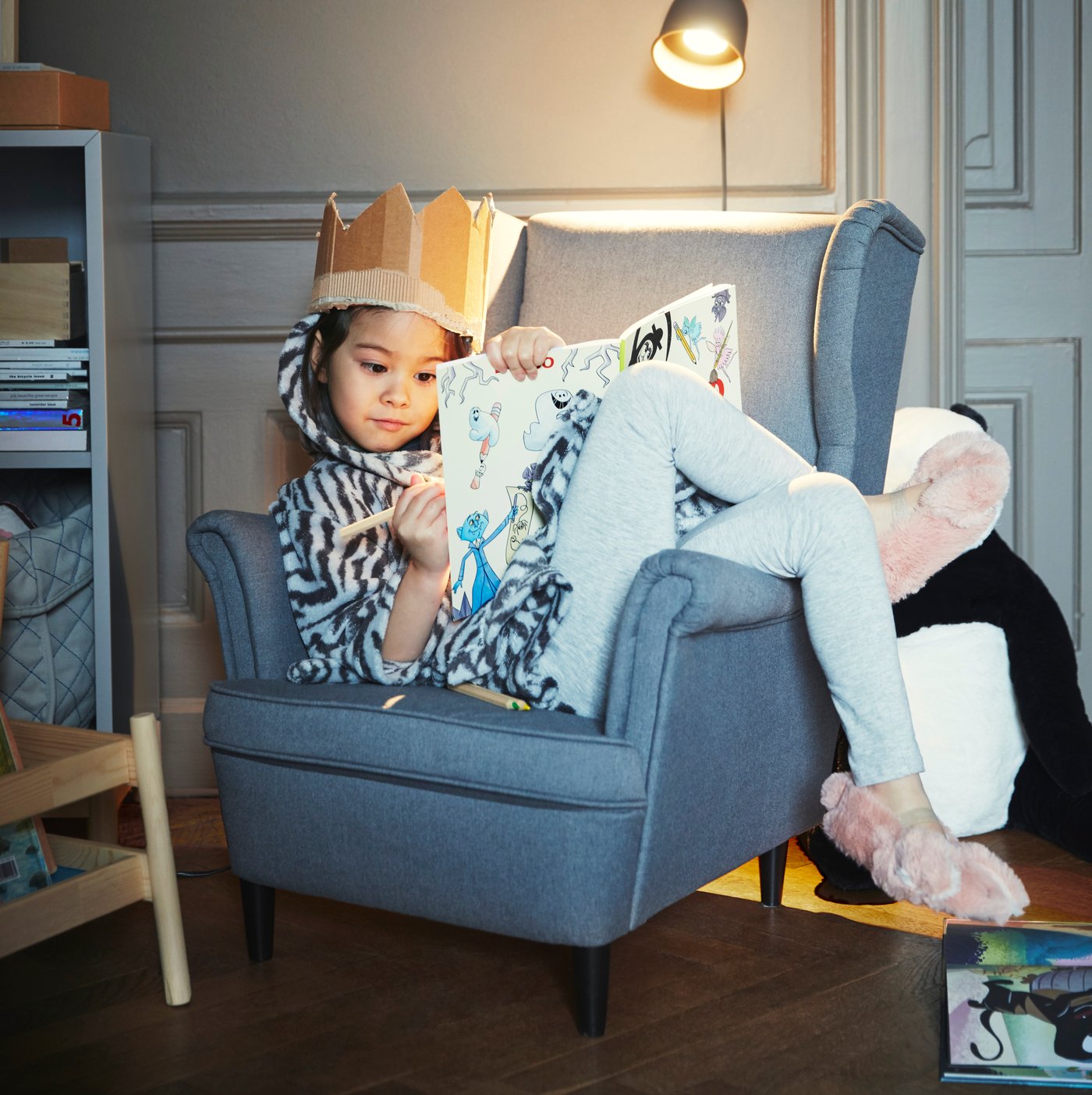 A girl wearing a crown is sitting in a grey STRANDMON children's armchair in the childrens room.